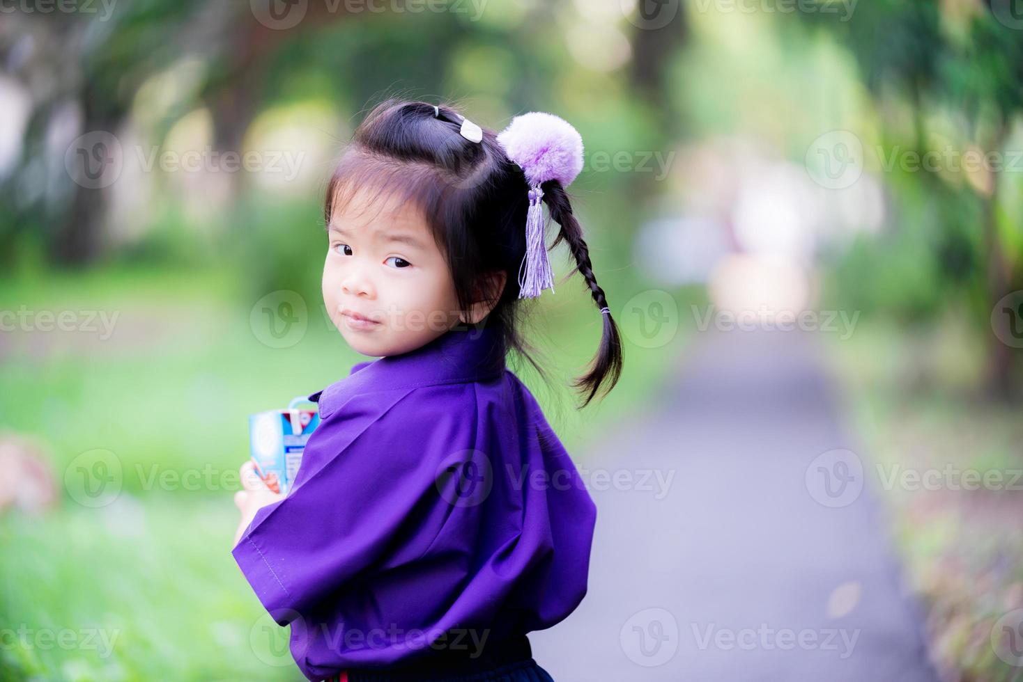 adorável garota em pé segurando a caixa de leite e se inclinou olhando para a câmera atrás dela. estudante vestindo uniforme escolar roxo. menina doce sorridente de 3 anos de idade. foto