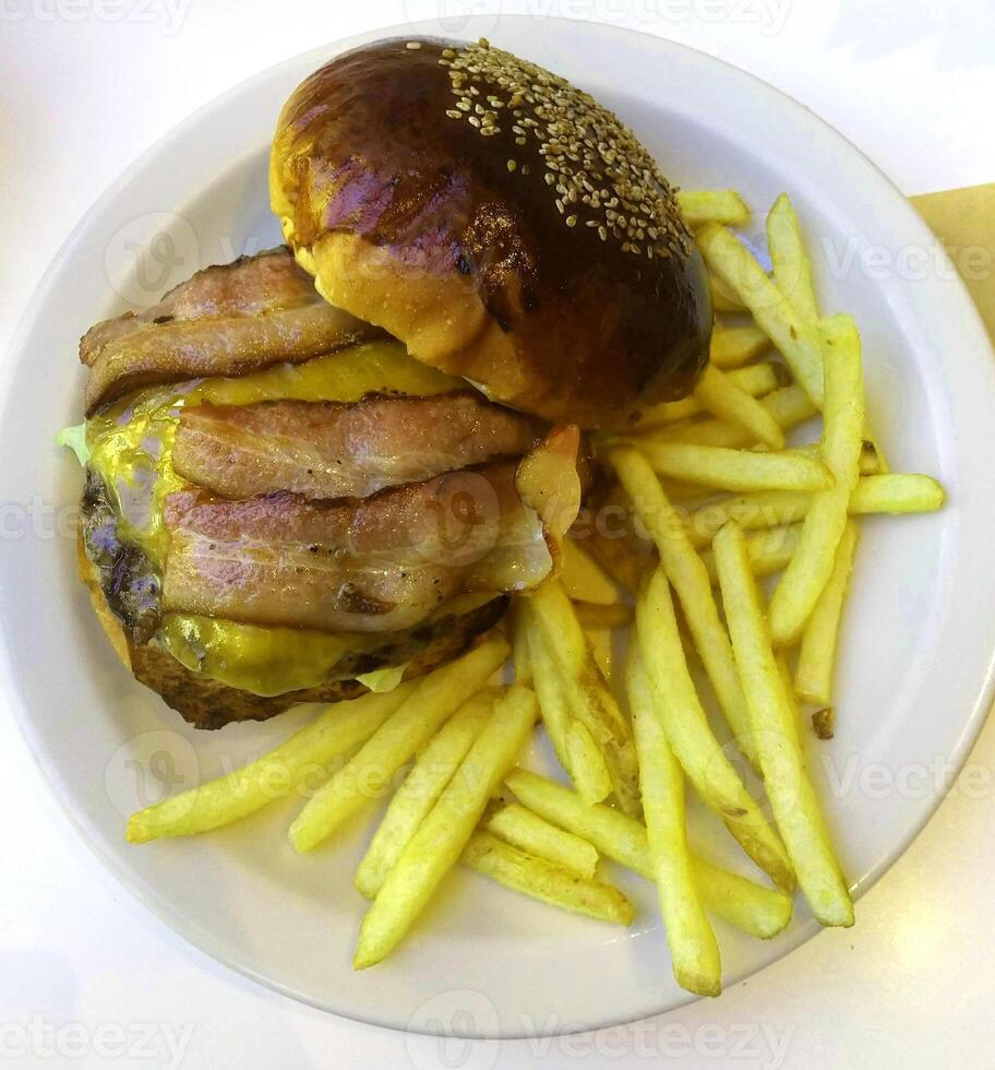 hambúrguer de carne fresca de comida de rua com pão integral, legumes, queijo e molhos e batatas fritas fritas no prato branco. foto