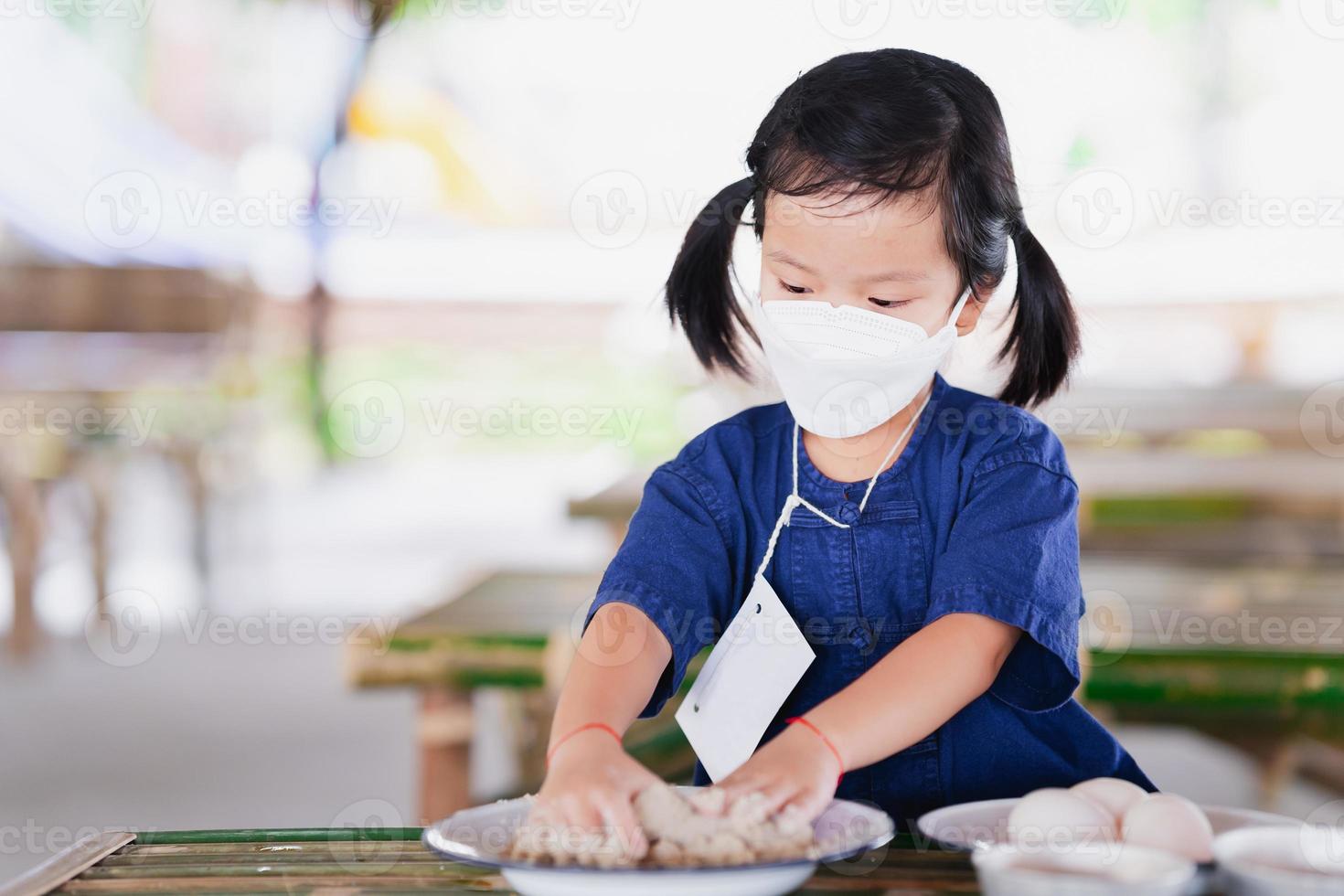 Menina de 4 anos usando máscara facial branca está usando duas mãozinhas. garoto mistura farinha branca, sal e água em um prato de zinco na mesa de bambu. criança aprendendo a preservar ovos salgados de ovos de pato. foto