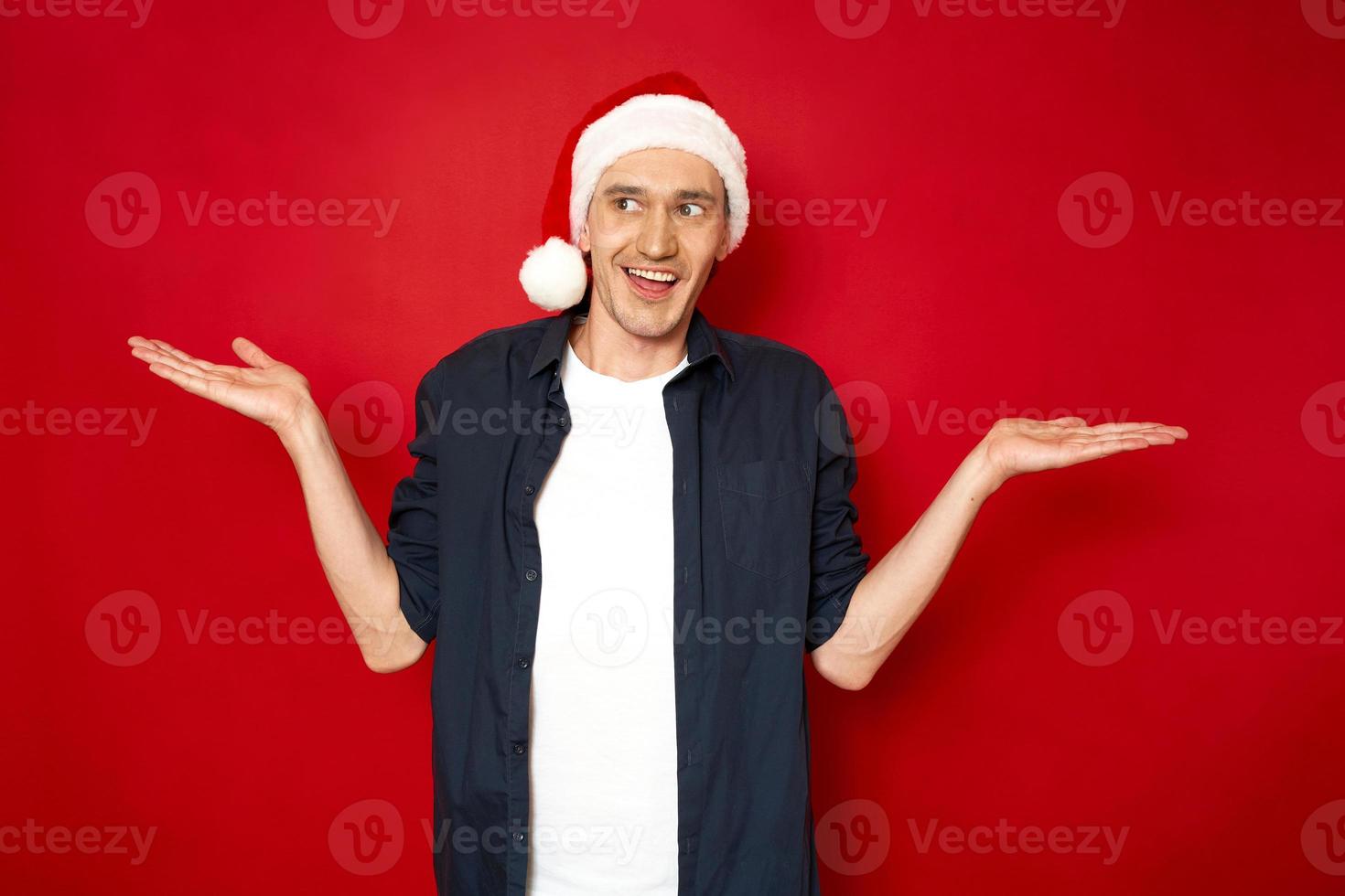 foto horizontal de natal de homem atraente com expressão perplexa espalha as mãos com olhar confuso, ele sorri absurdamente, usa roupas casuais e sente-se hesitante isolado sobre fundo vermelho. conceito de dúvida