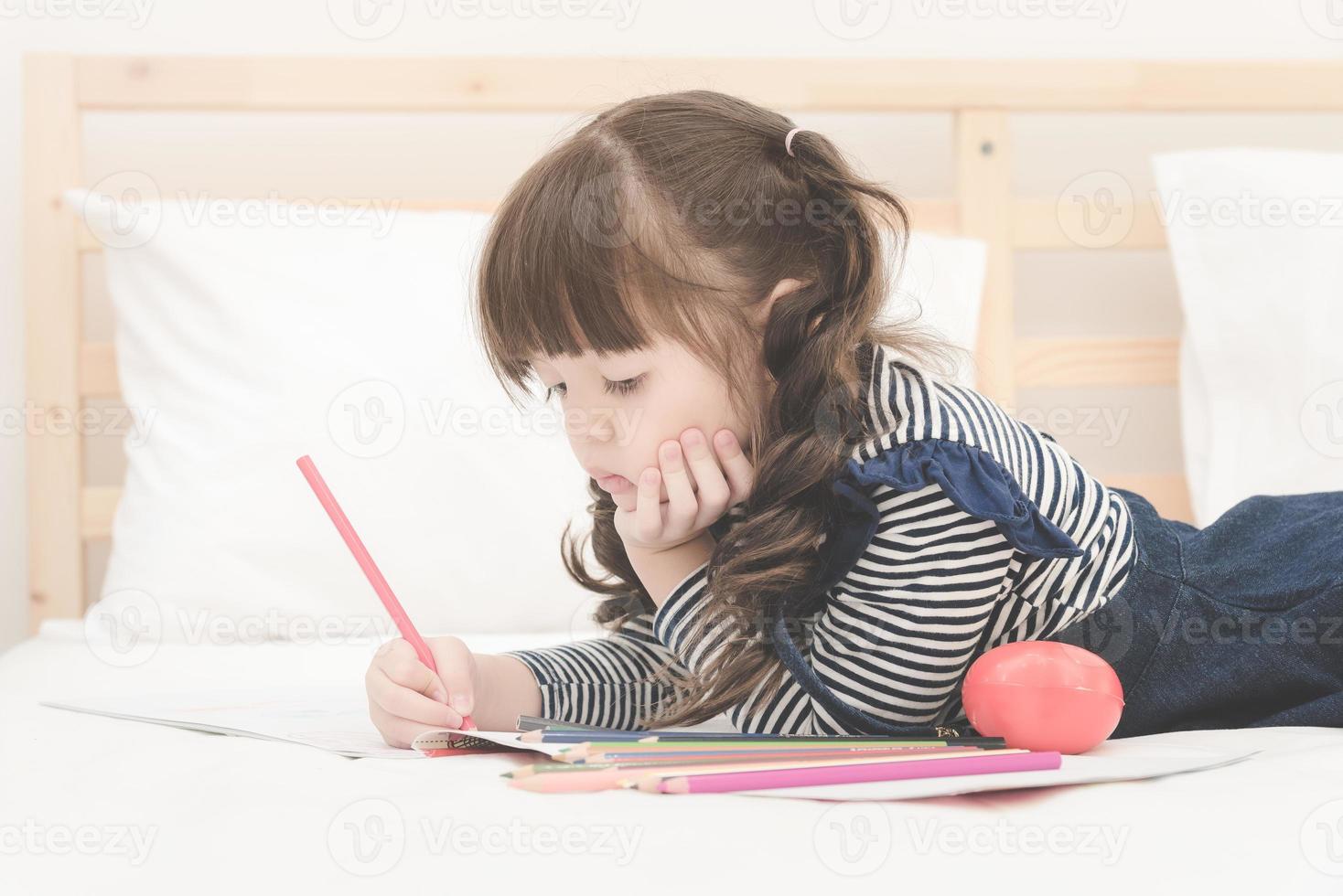 menina asiática bonitinha fazendo lição de casa, escrevendo com lápis coloridos na cama em casa. design elegante para criança brincando, aprendizagem pré-escolar e conceito de educação de arte criativa. foto