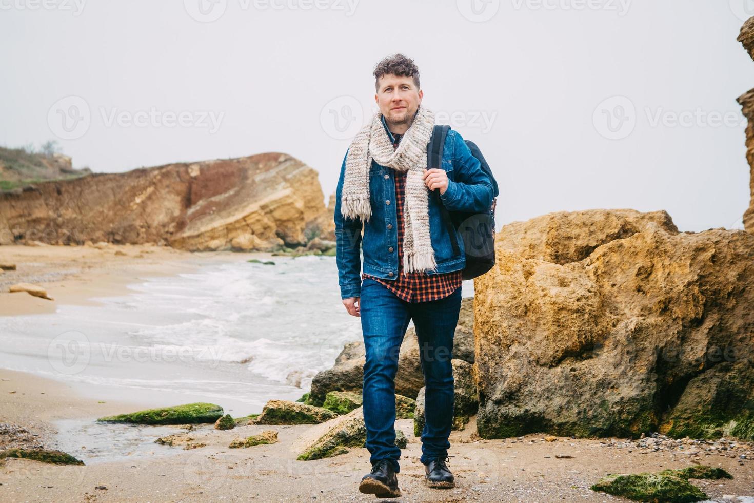 homem viajante com mochila em uma praia arenosa no meio de rochas contra o fundo do mar foto
