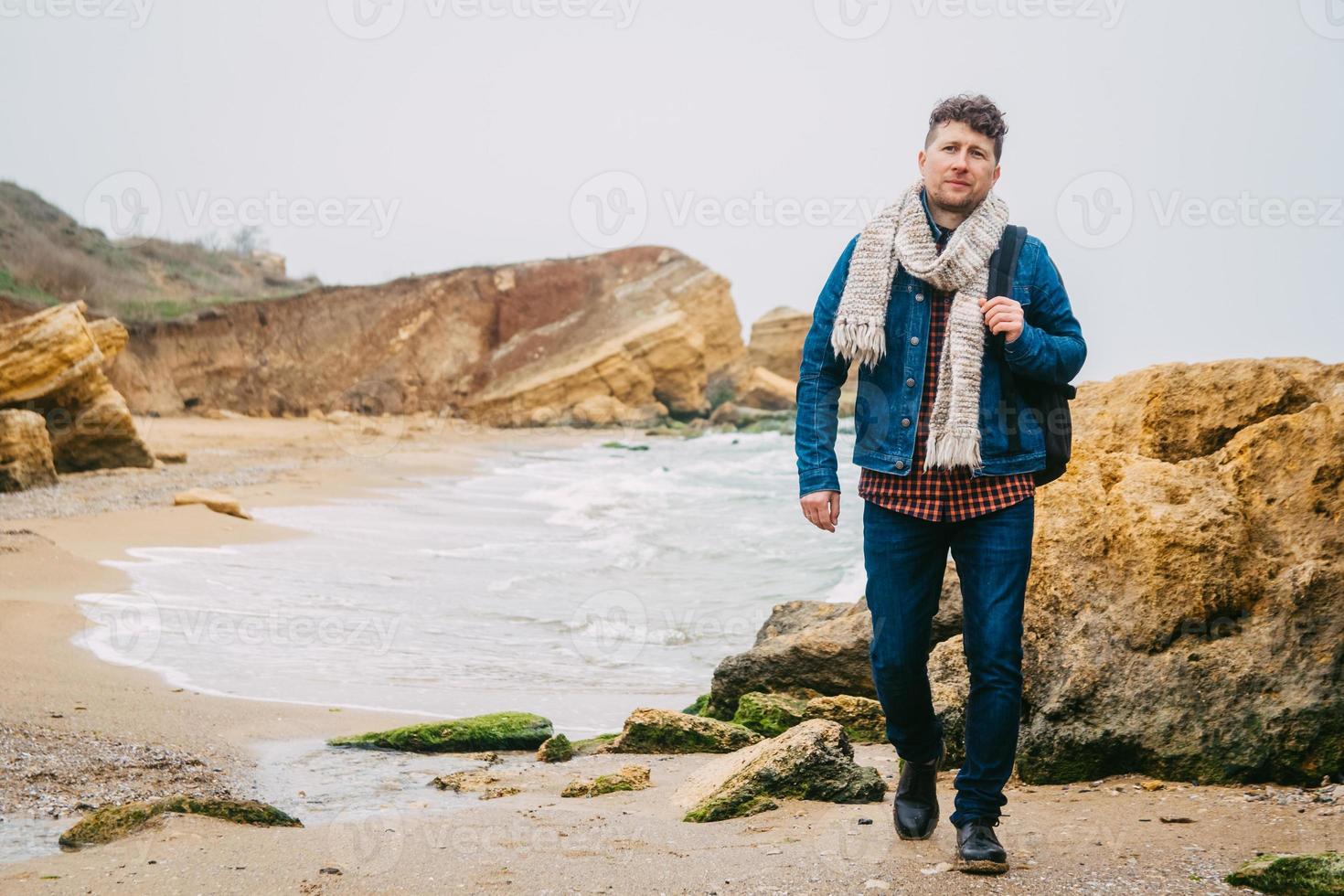 homem viajante com mochila em uma praia arenosa no meio de rochas contra o fundo do mar foto