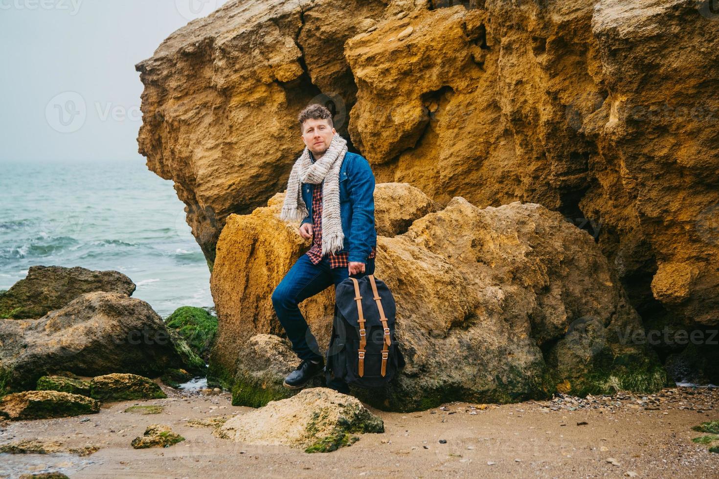 homem viajante com mochila em uma praia arenosa no meio de rochas contra o fundo do mar foto