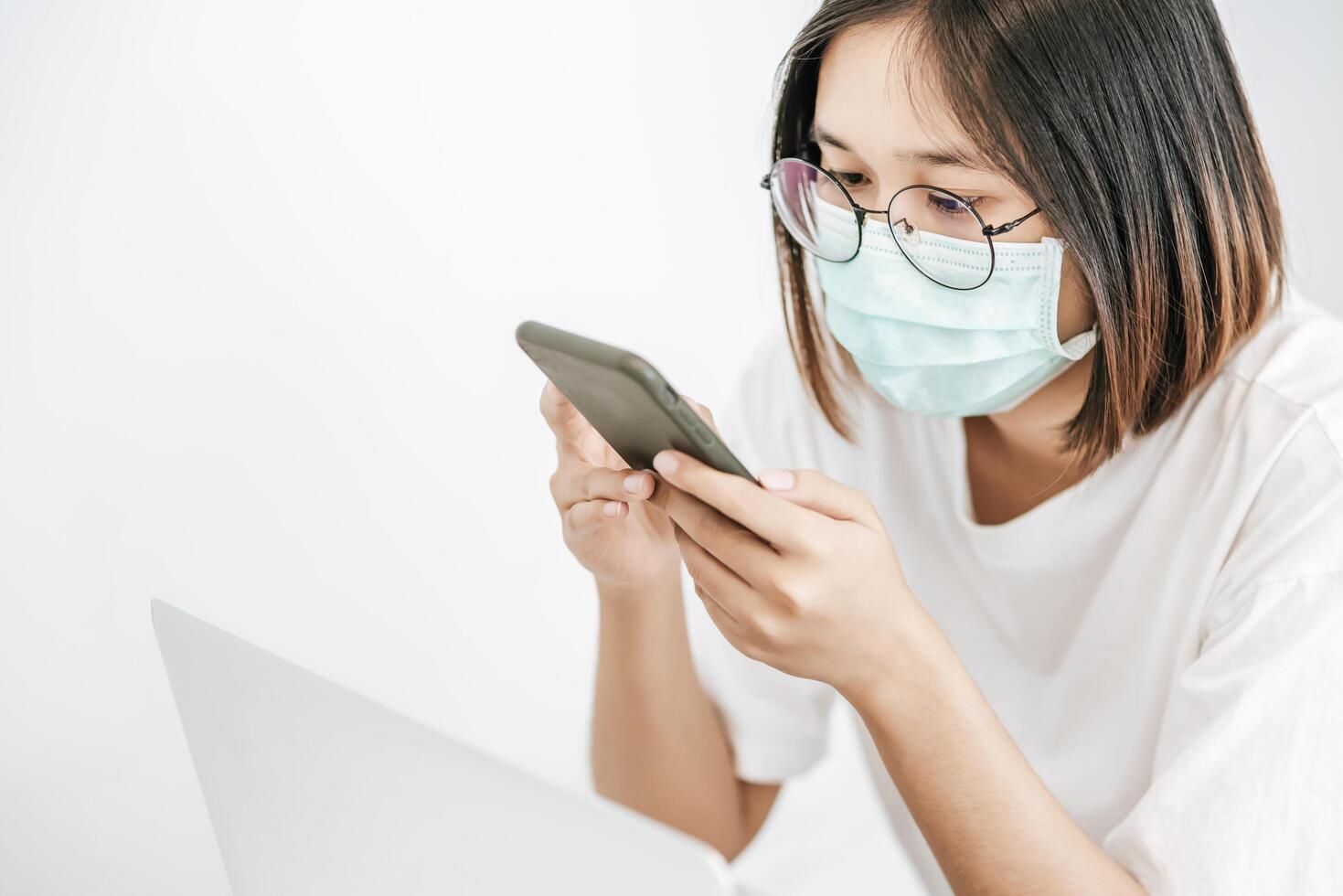 mulher usando uma máscara sanitária, jogando um smartphone e tendo um laptop. foto