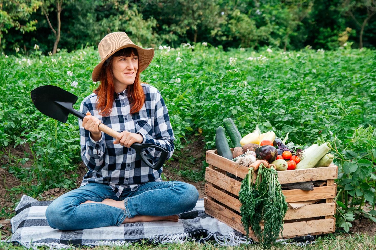 agricultora com uma pá sentada perto de vegetais orgânicos frescos em uma caixa de madeira contra o fundo de uma horta foto