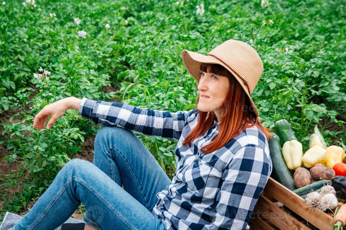 agricultora sentada em um cobertor perto de vegetais orgânicos frescos em uma caixa de madeira contra o fundo de uma horta foto