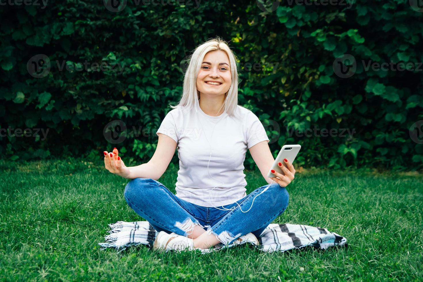 mulher com fones de ouvido e smartphone nas mãos sentada em pose meditativa ouvindo música na grama verde foto