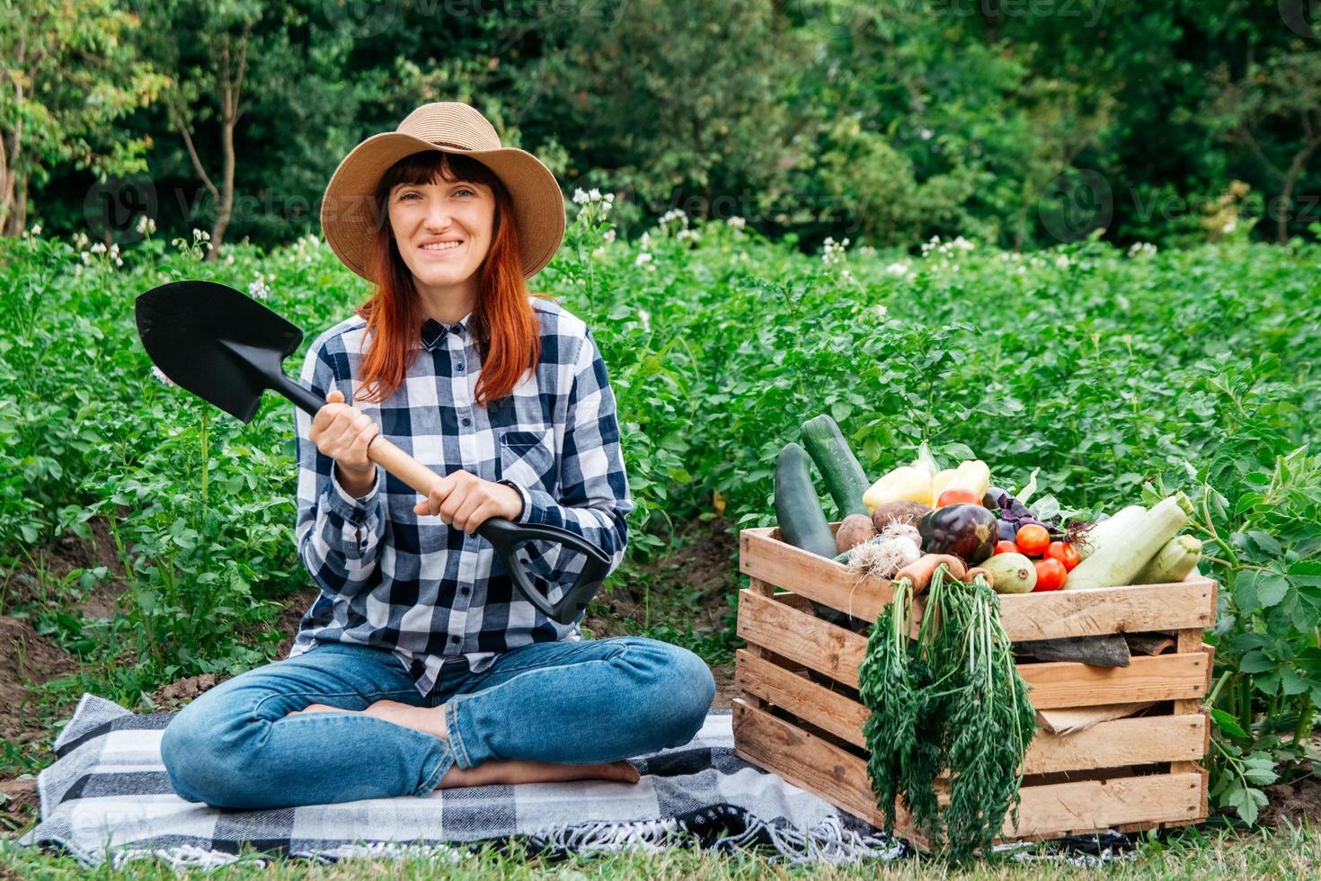 agricultora com uma pá sentada perto de vegetais orgânicos frescos em uma caixa de madeira contra o fundo de uma horta foto