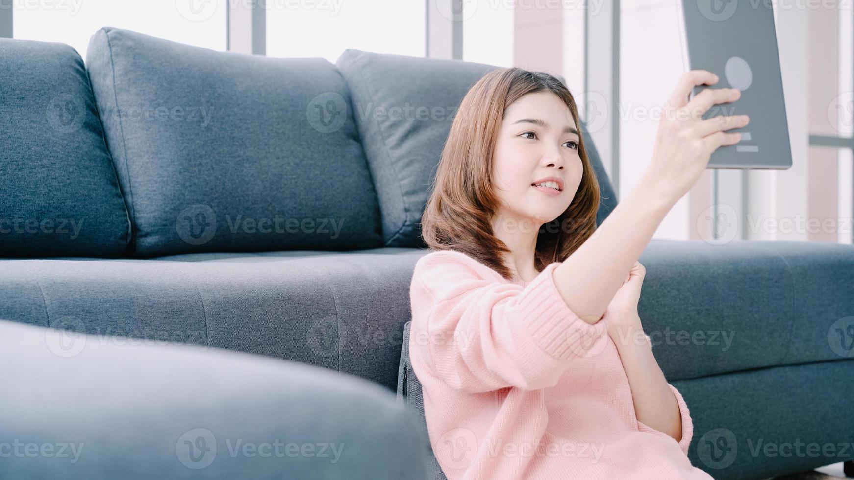 mulher asiática usando tablet enquanto estava deitada no sofá em sua sala de estar. feliz feminino usar tablet para videochamada com os amigos ou namorado em casa. foto