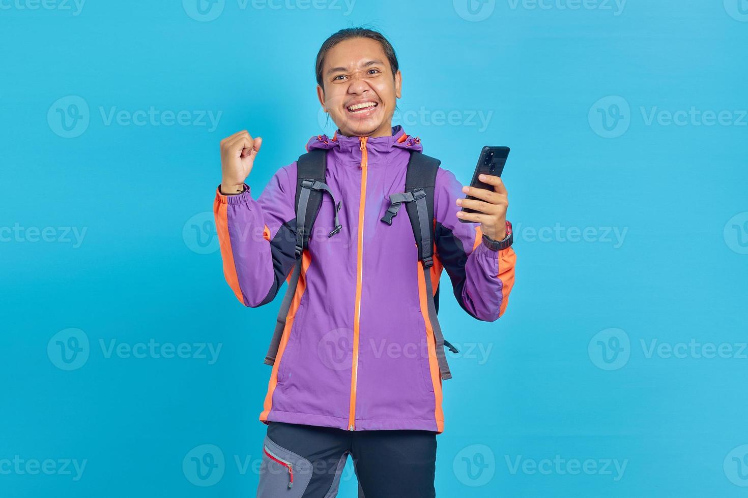 retrato de jovem asiático animado comemorando com telefone celular isolado em fundo azul foto