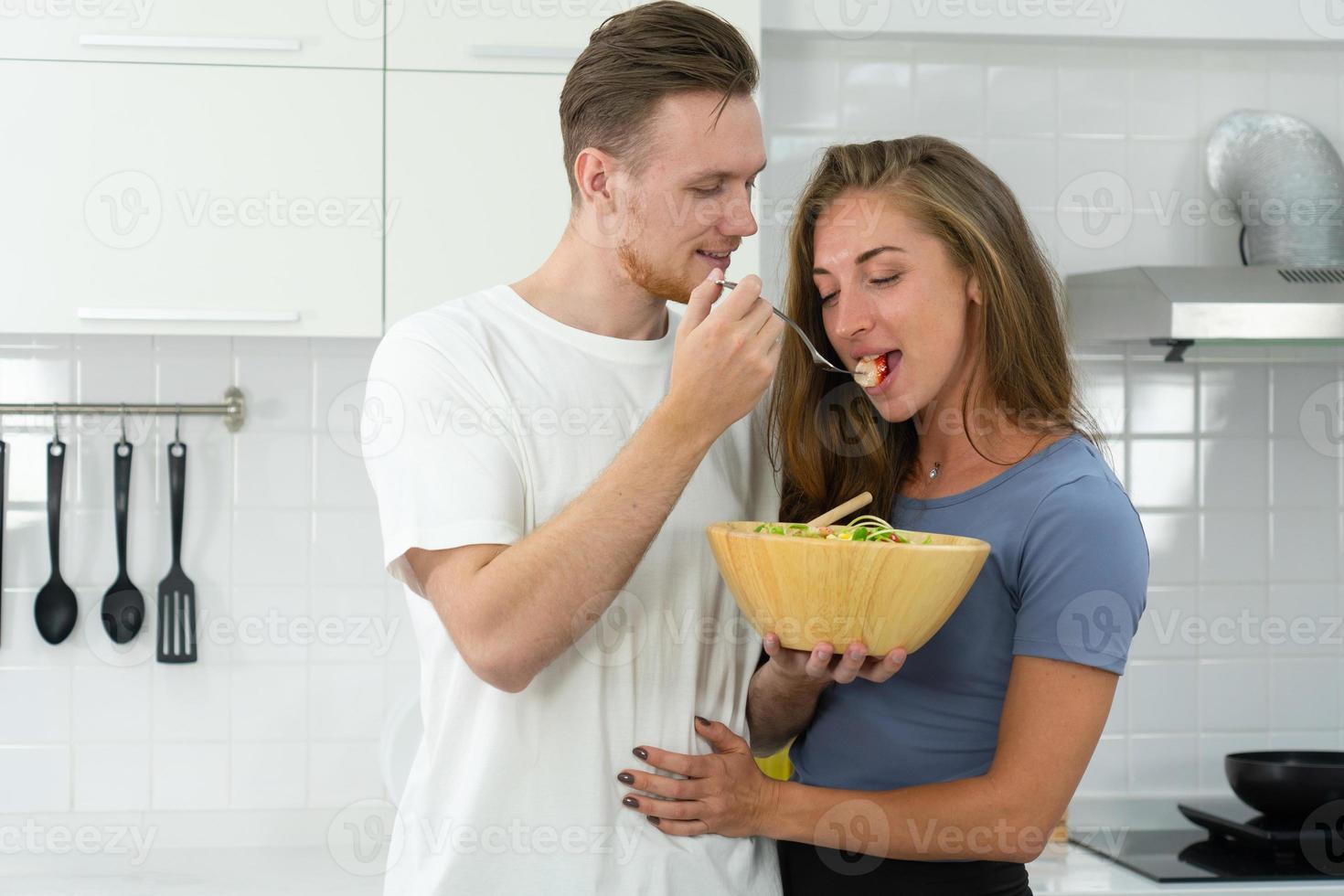 jovem namorado caucasiano alimentando a salada de frutas orgânicas saudáveis no café da manhã com a namorada na cozinha. conceito casal junto foto