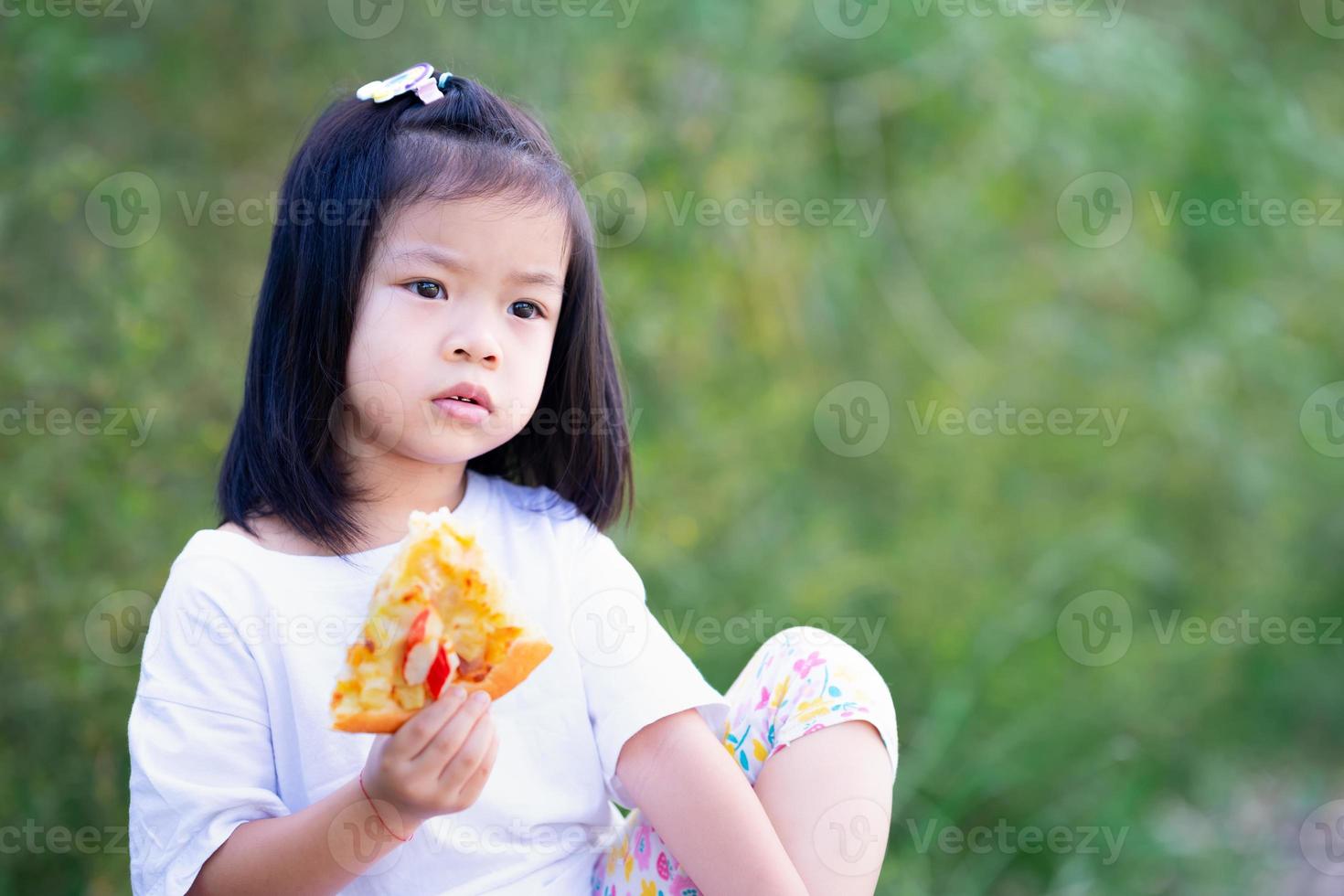 menina criança sentou-se comendo pizza e olhou para a distância. espaço vazio para inserir texto. criança tem 4 anos. bebê vestindo t-shirt branco. foto