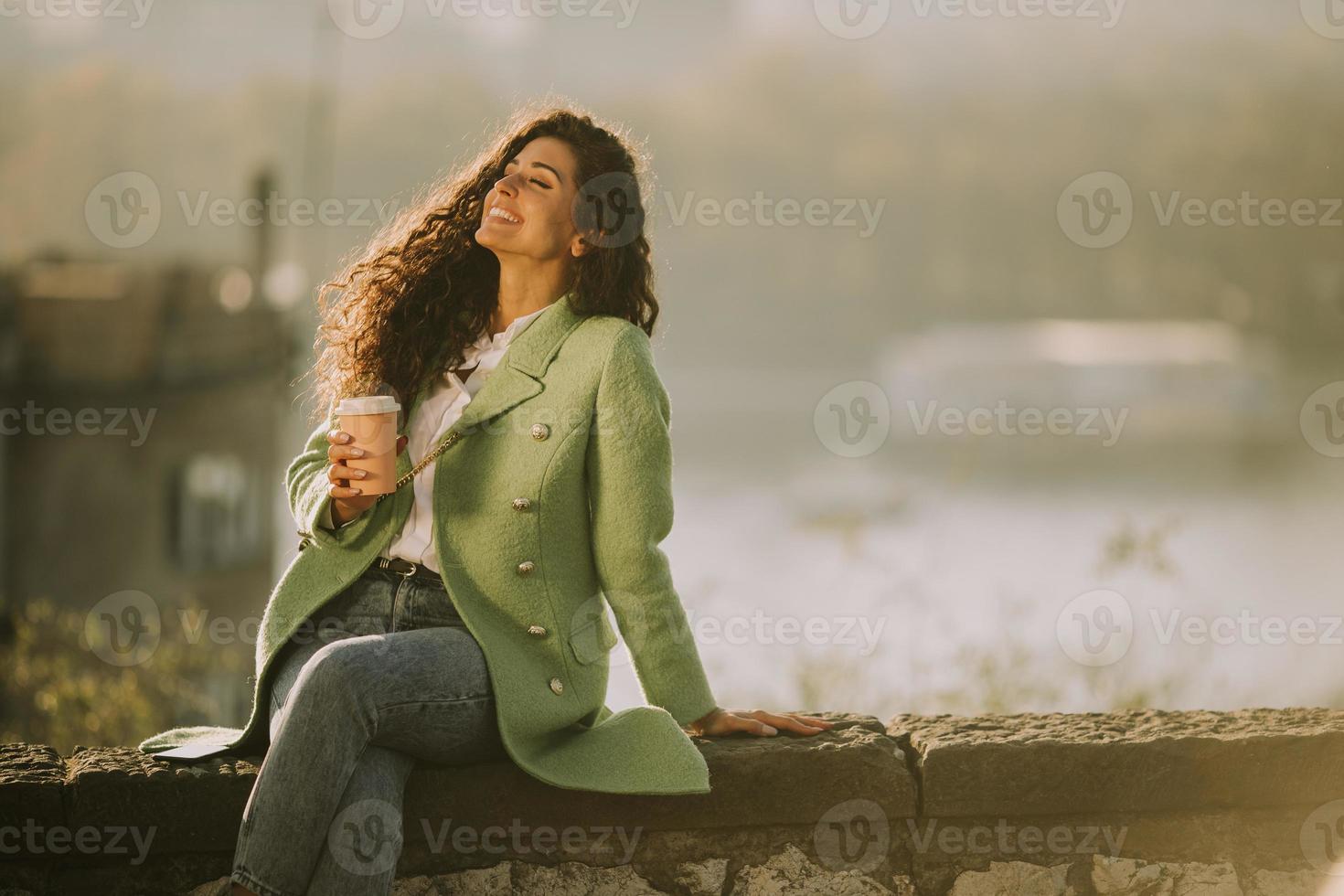 bela jovem curtindo o sol de outono enquanto está sentada perto do rio e bebendo café para viagem foto