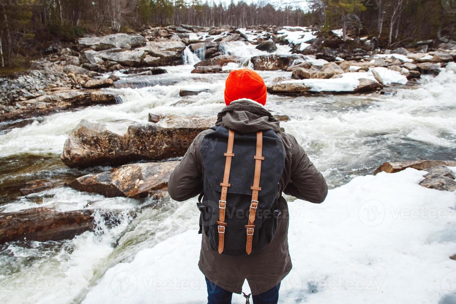 homem viajante com uma mochila de pé sobre uma rocha no rio de montanha e a cachoeira que flui entre as rochas cobertas de neve e a floresta. atirar pelas costas foto