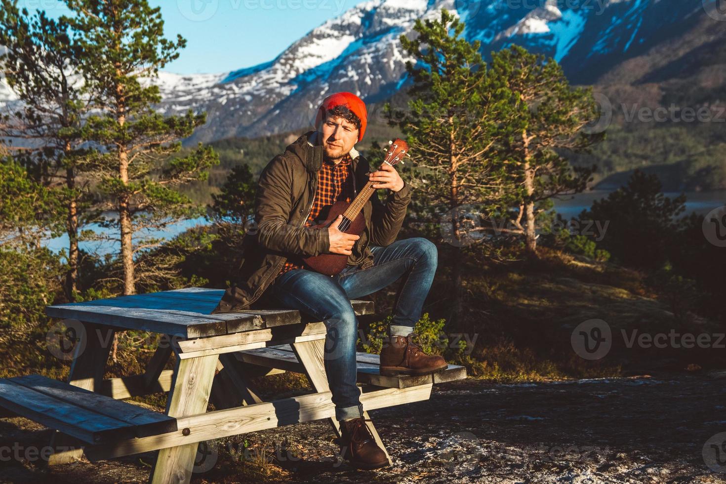 jovem tocando violão, sentado em uma mesa de madeira no contexto de montanhas, florestas e lagos. relaxando e curtindo os dias de sol. lugar para texto ou publicidade foto