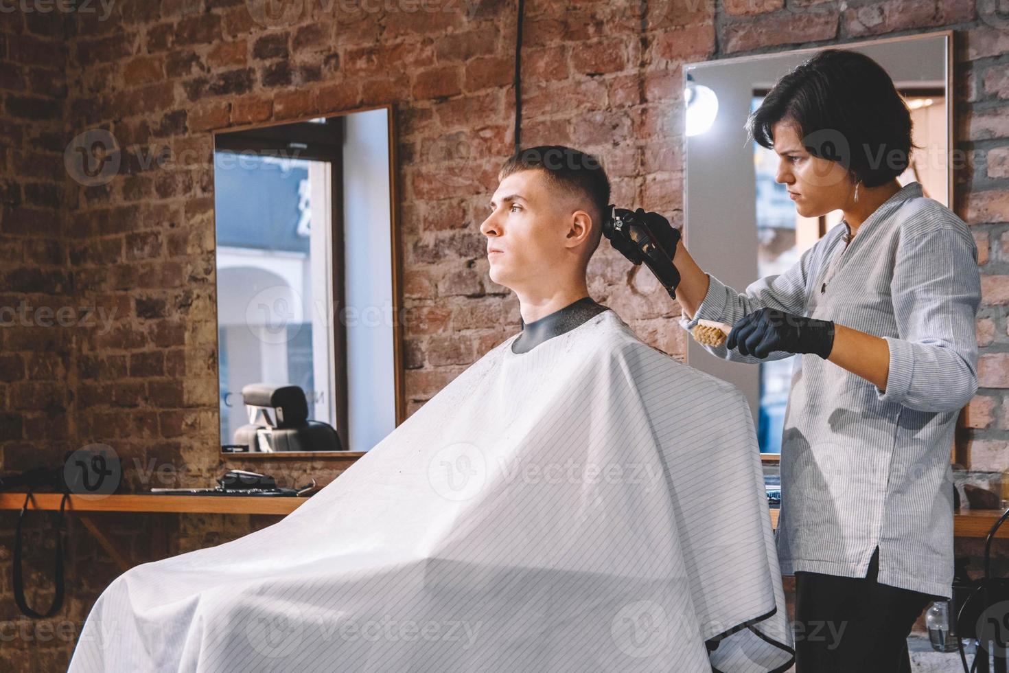 cabeleireira linda corta o cabelo da cabeça da cliente com um aparador elétrico na barbearia. publicidade e conceito de barbearia. lugar para texto ou publicidade foto