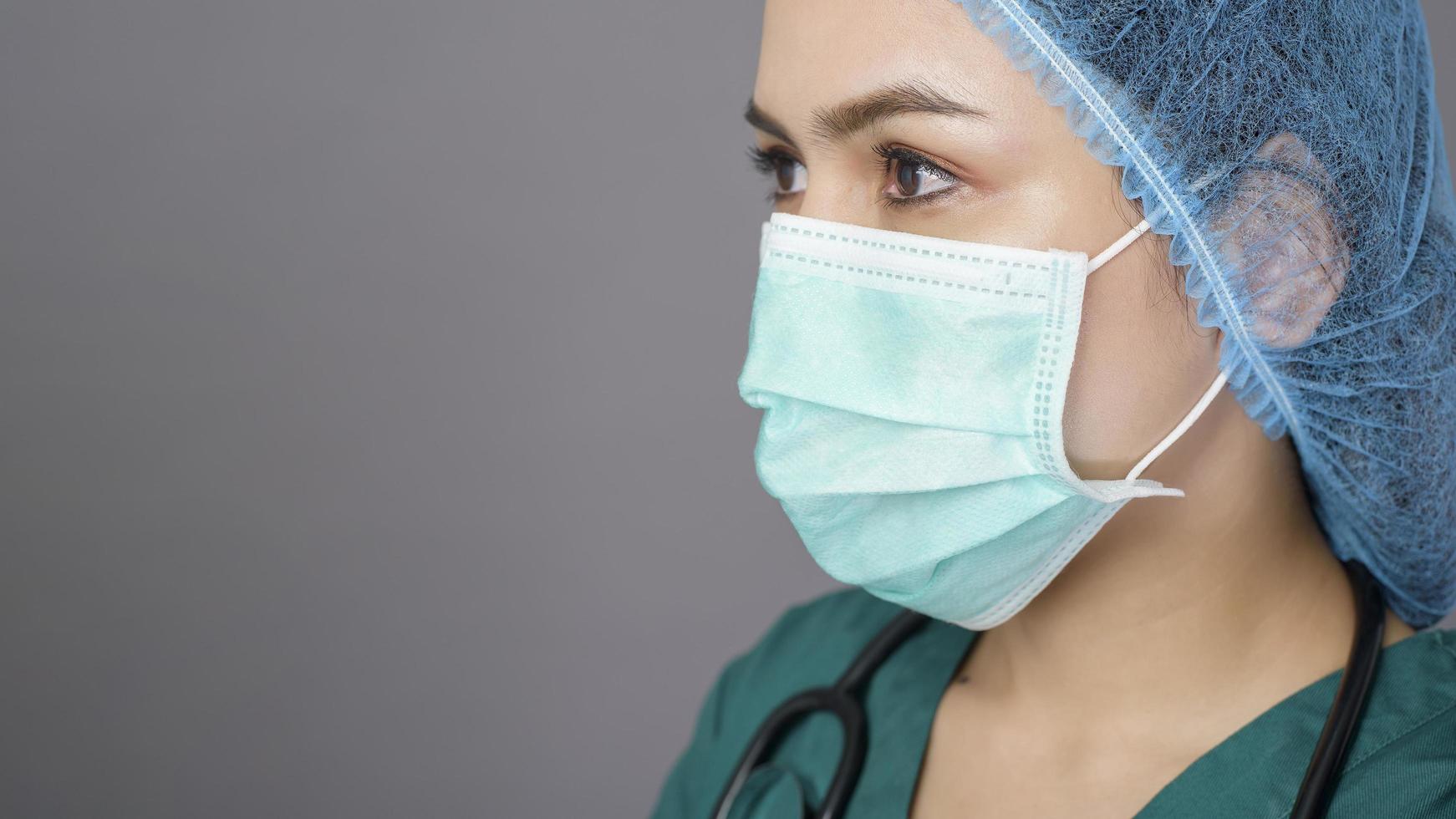 jovem médica confiante em uniforme verde está usando máscara cirúrgica sobre estúdio de fundo cinza foto