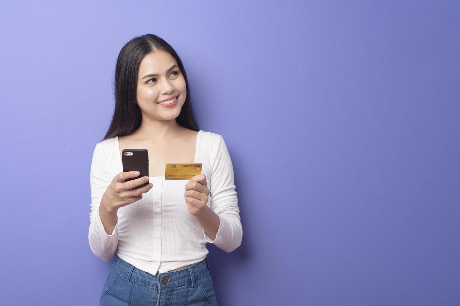retrato de jovem asiática está usando telefone celular com cartão de crédito em fundo roxo foto