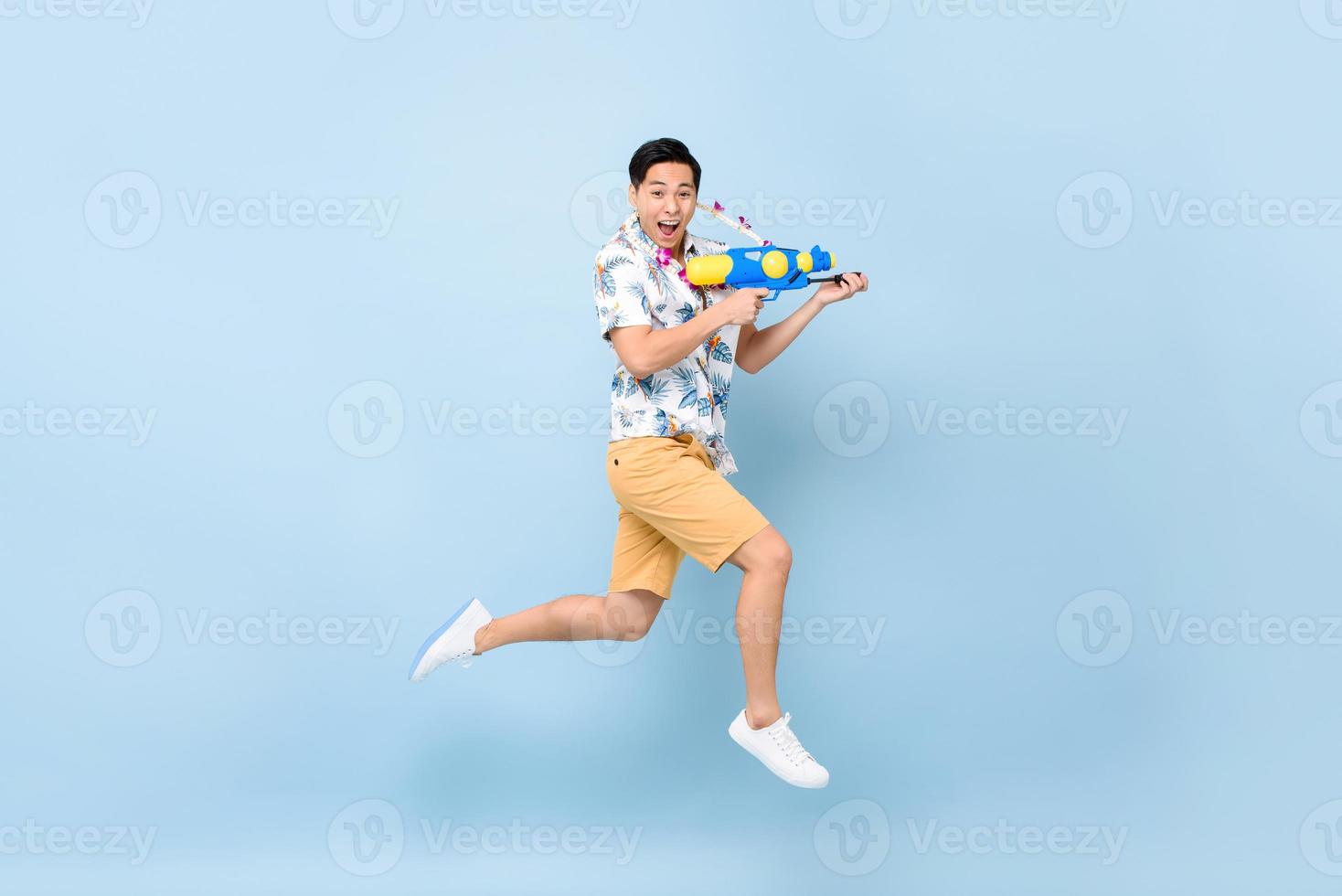 bonito sorridente jovem asiático brincando com pistola d'água e pulando em fundo azul estúdio para festival songkran na tailândia e sudeste da ásia foto