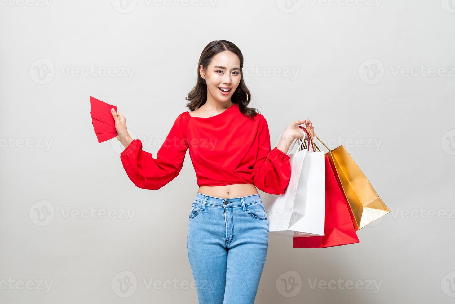 feliz mulher asiática segurando sacolas de compras e envelopes vermelhos isolados no fundo cinza do estúdio para o conceito de venda de ano novo chinês foto