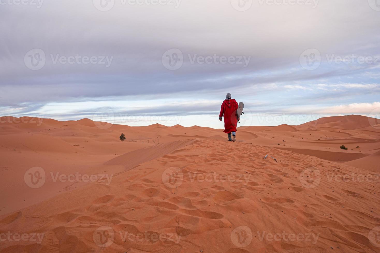 homem em roupas tradicionais com sandboard andando nas dunas de areia contra o céu foto