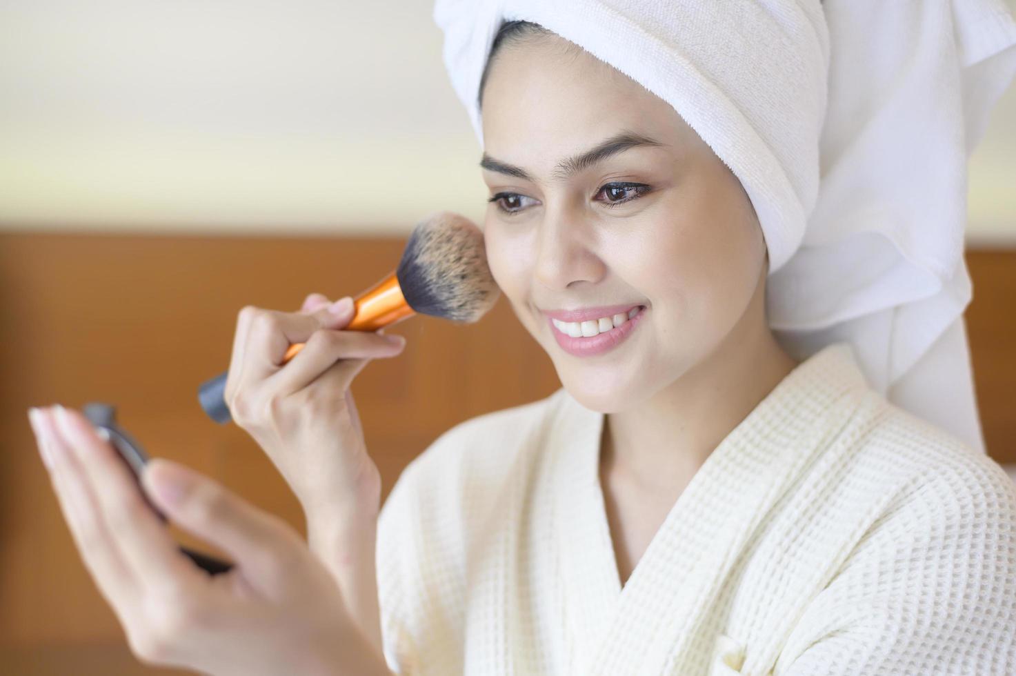 mulher feliz atraente em roupão branco está aplicando maquiagem natural com pincel de pó cosmético, conceito de beleza. foto