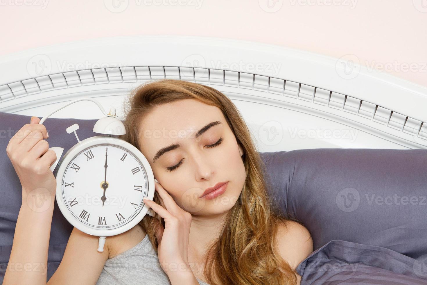 conceito de trabalho dormido demais, insônia de sono ruim - jovem linda garota sonolenta com os olhos fechados segura um relógio e deita-se em sua cama no quarto de manhã foto