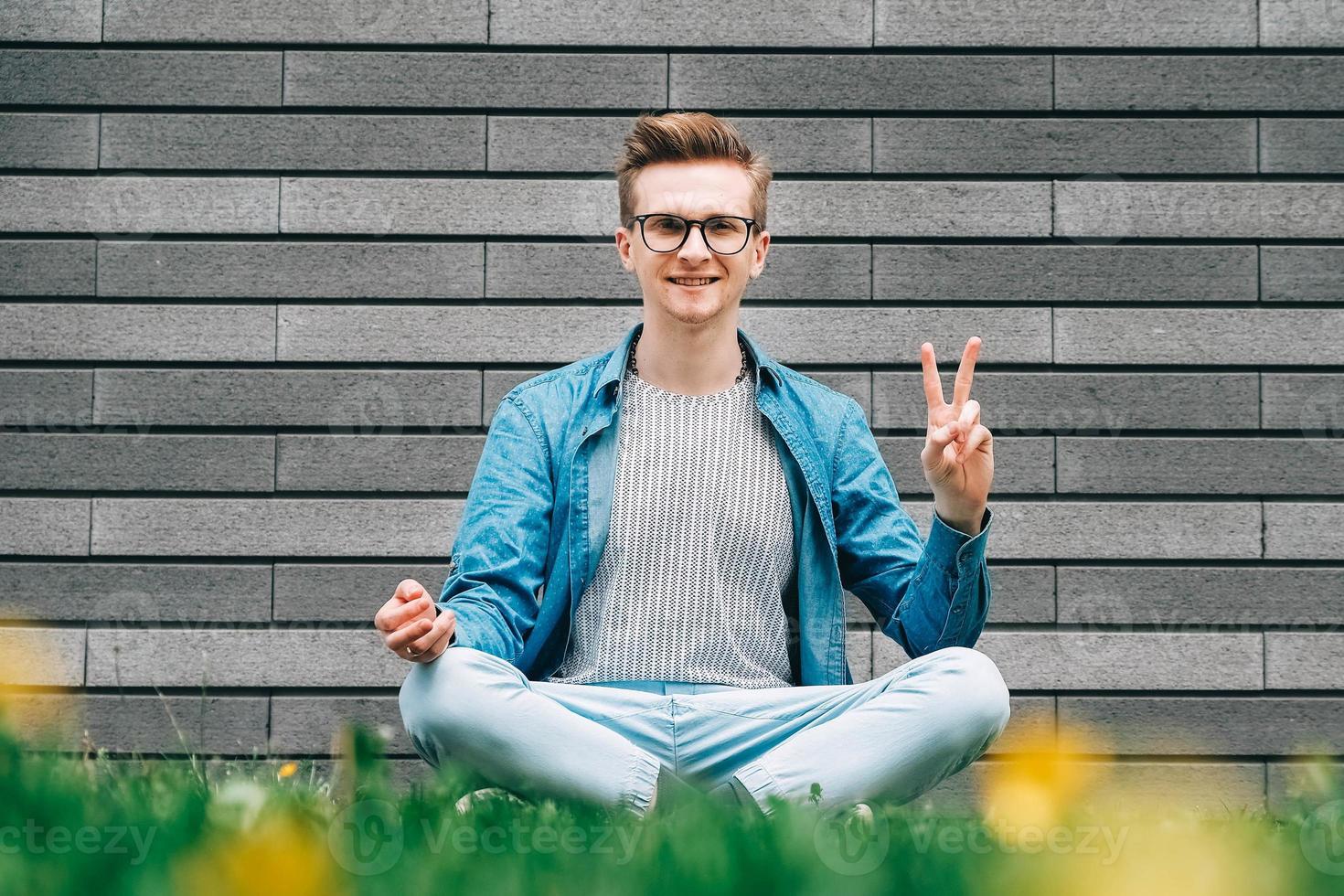 homem de camisa, jeans e óculos sentado na pose de meditação na grama verde em um fundo de uma parede cinza foto