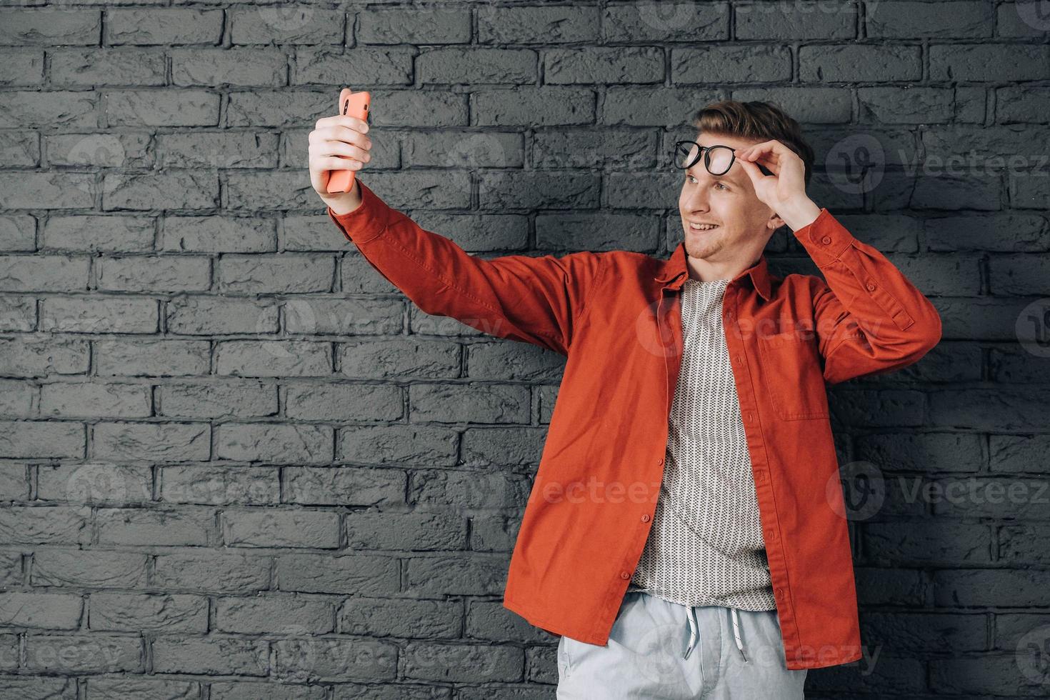 jovem alegre de camisa vermelha e óculos tirando foto de selfie no celular em um fundo de parede de tijolo preto. copiar, espaço vazio para texto