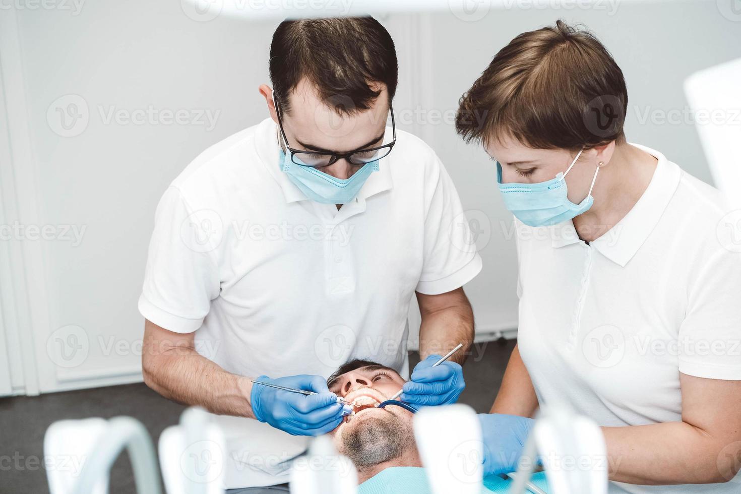 dentista e seu assistente tratam um paciente do sexo masculino em uma clínica odontológica. dentistas em máscaras médicas trabalhando com os dentes do cliente foto