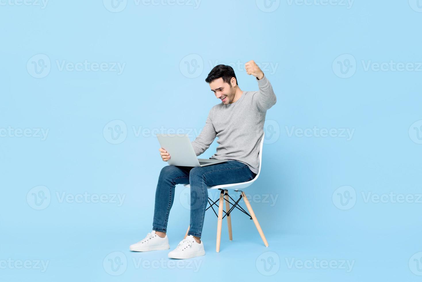 surpreso jovem homem caucasiano olhando para o computador portátil e levantando o punho fazendo sim gesto isolado em fundo azul claro foto