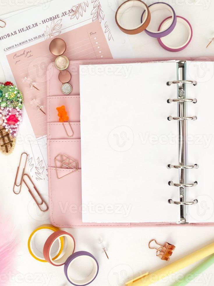 planejador com páginas abertas em um fundo branco e com lindos acessórios canetas, botões, alfinetes e fita colorida. vista superior de um planejador rosa com artigos de papelaria de negócios foto