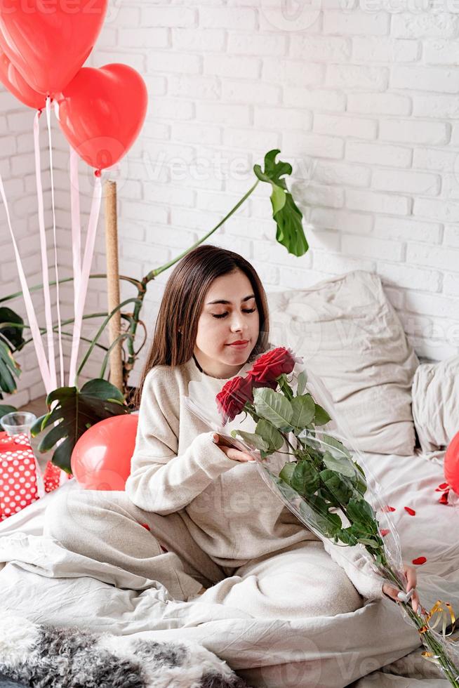 jovem mulher bonita sentada na cama comemorando o dia dos namorados segurando rosas vermelhas foto