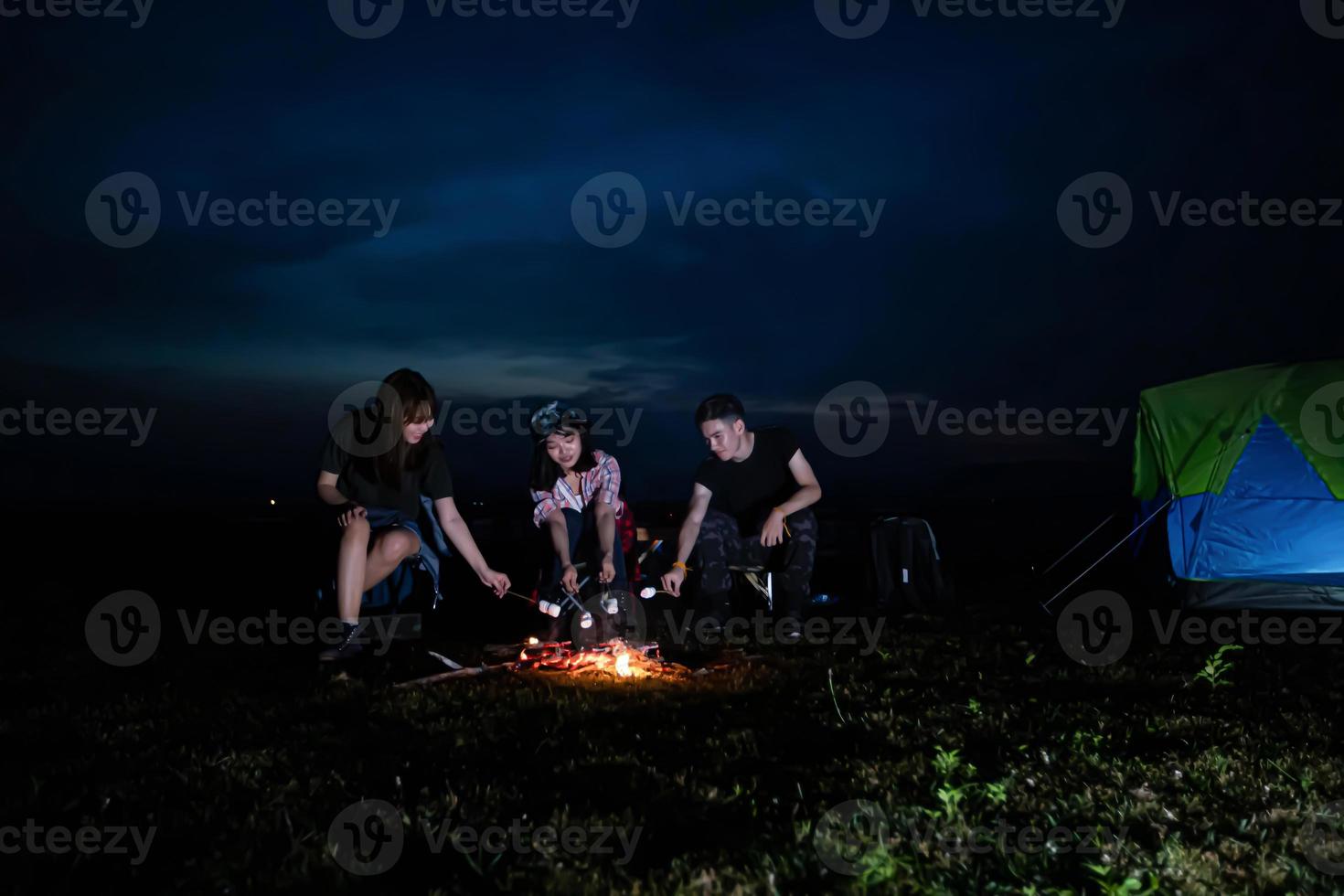 um grupo de amigos asiáticos turista bebendo cerveja alcoólica e tocando violão junto com a felicidade no verão enquanto acampa e churrasco marshmallows em espetos sobre fogueira perto do lago foto