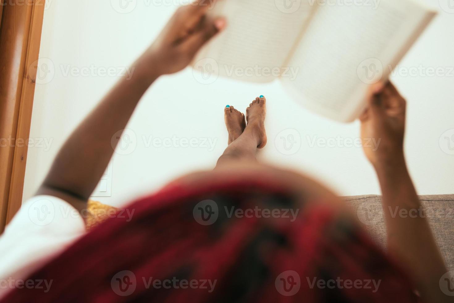 mulher negra deitada no sofá lendo um livro com os pés na parede foto