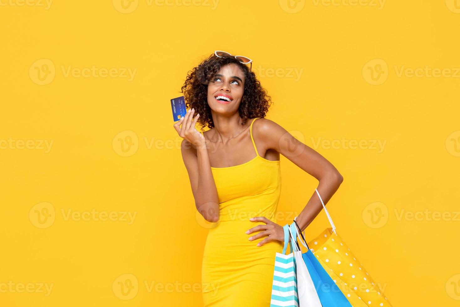 retrato de mulher afro-americana atraente pensando enquanto carregava sacolas de compras e segurando seu cartão de crédito em fundo amarelo estúdio isolado foto