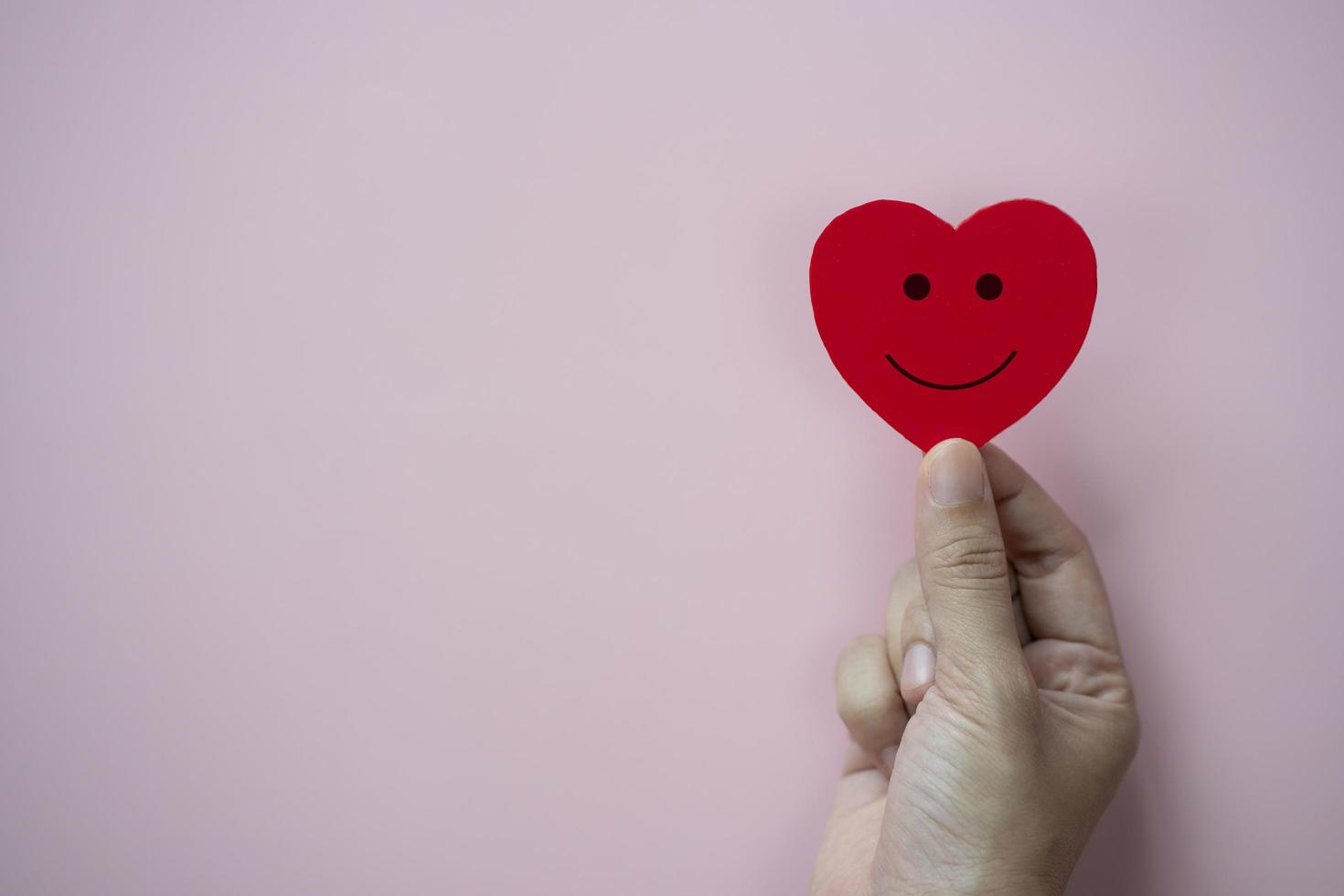 mãos segurando um coração vermelho com ícone de rosto sorridente em fundo rosa pastel. foto