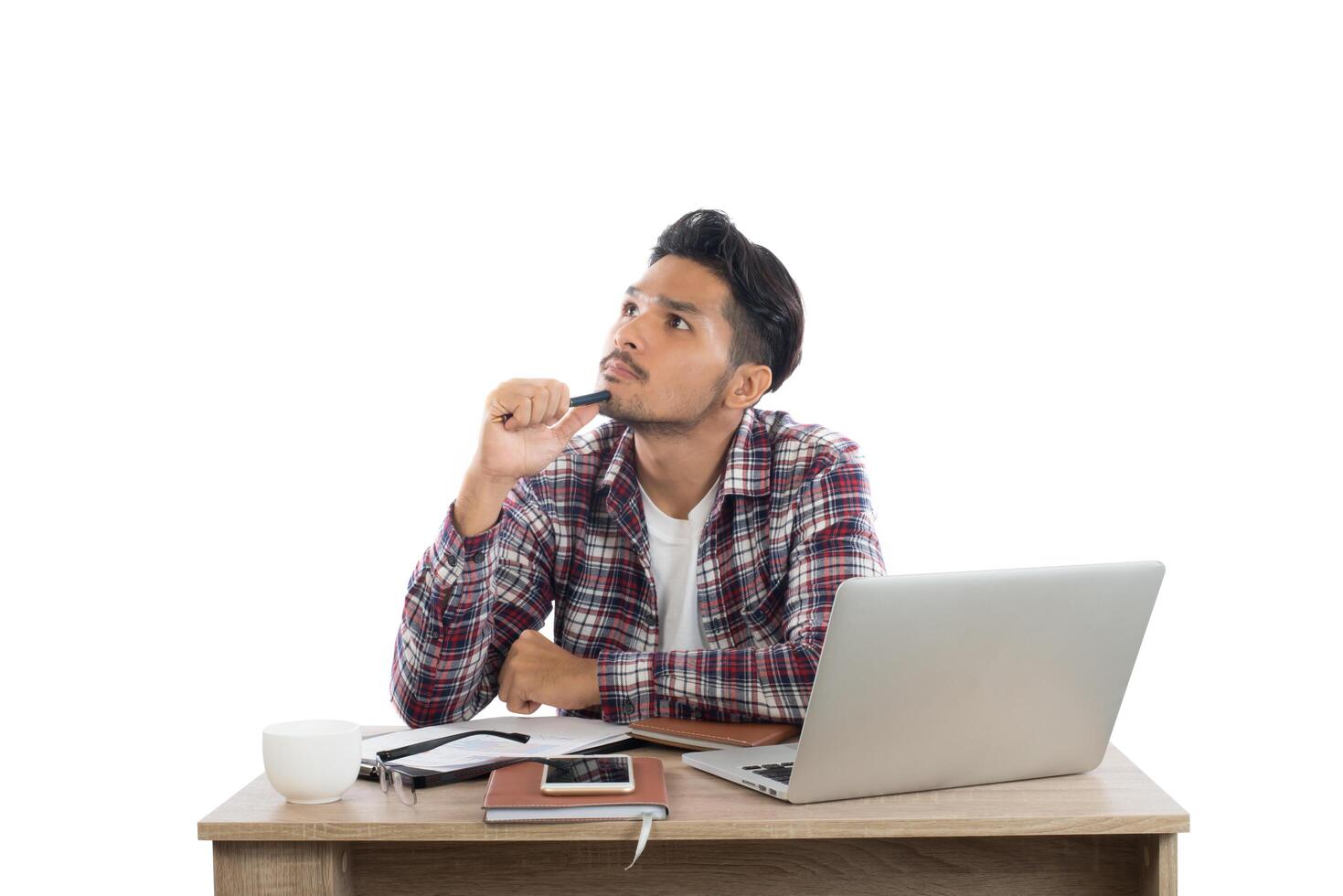 pensativo jovem segurando a mão no queixo e desviar o olhar enquanto está sentado em seu local de trabalho isolado no fundo branco. foto