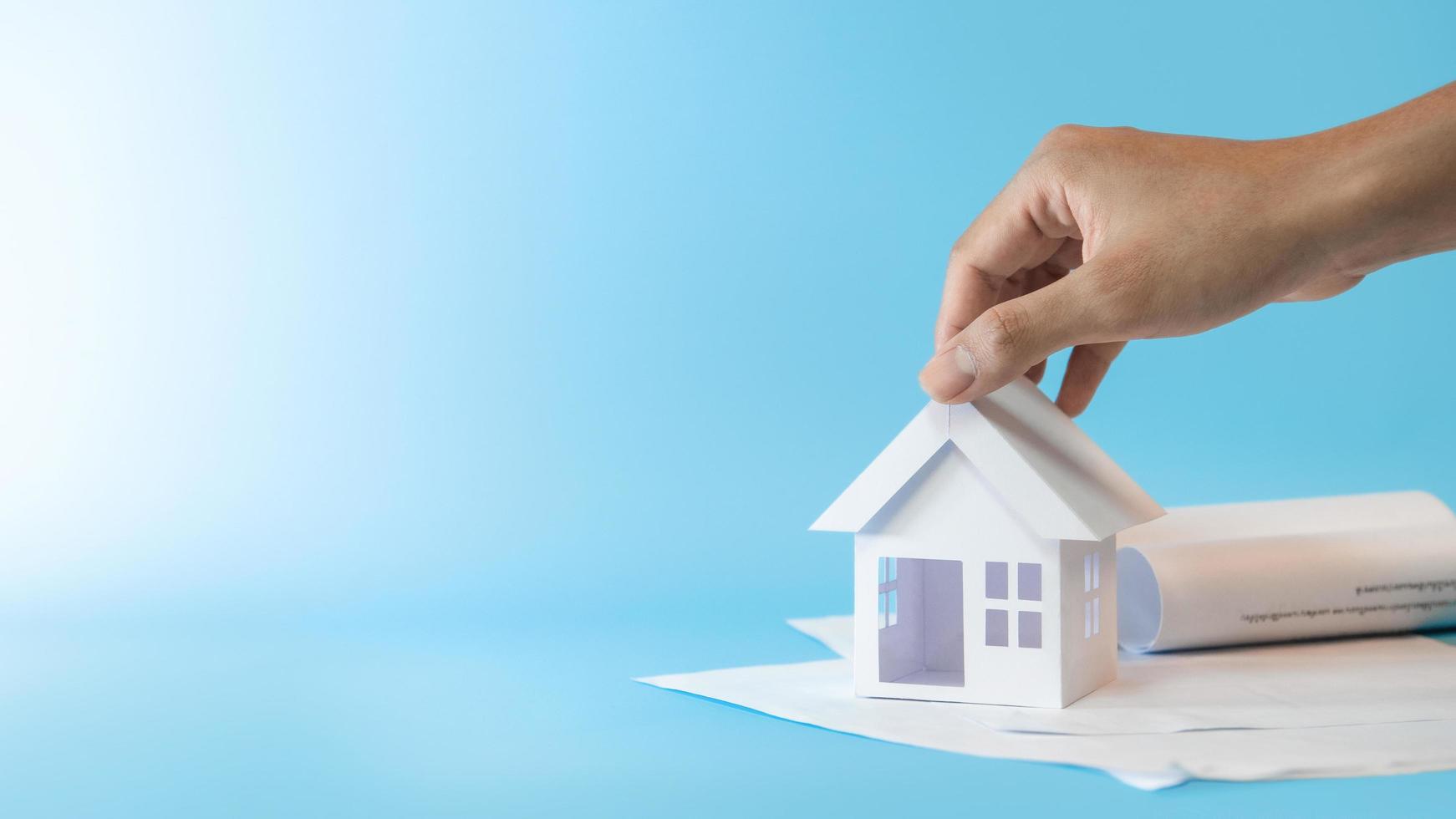 mão de homem tocando o modelo de casa branca em documentos comerciais em fundo azul para plano de refinanciamento e conceito imobiliário. foto
