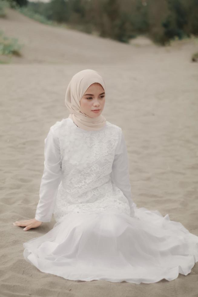 linda modelo feminina islâmica vestindo moda hijab, um vestido de noiva moderno para mulher muçulmana sentada na areia e na praia. retrato um modelo de menina asiática usando hijab se divertindo na praia com árvores foto