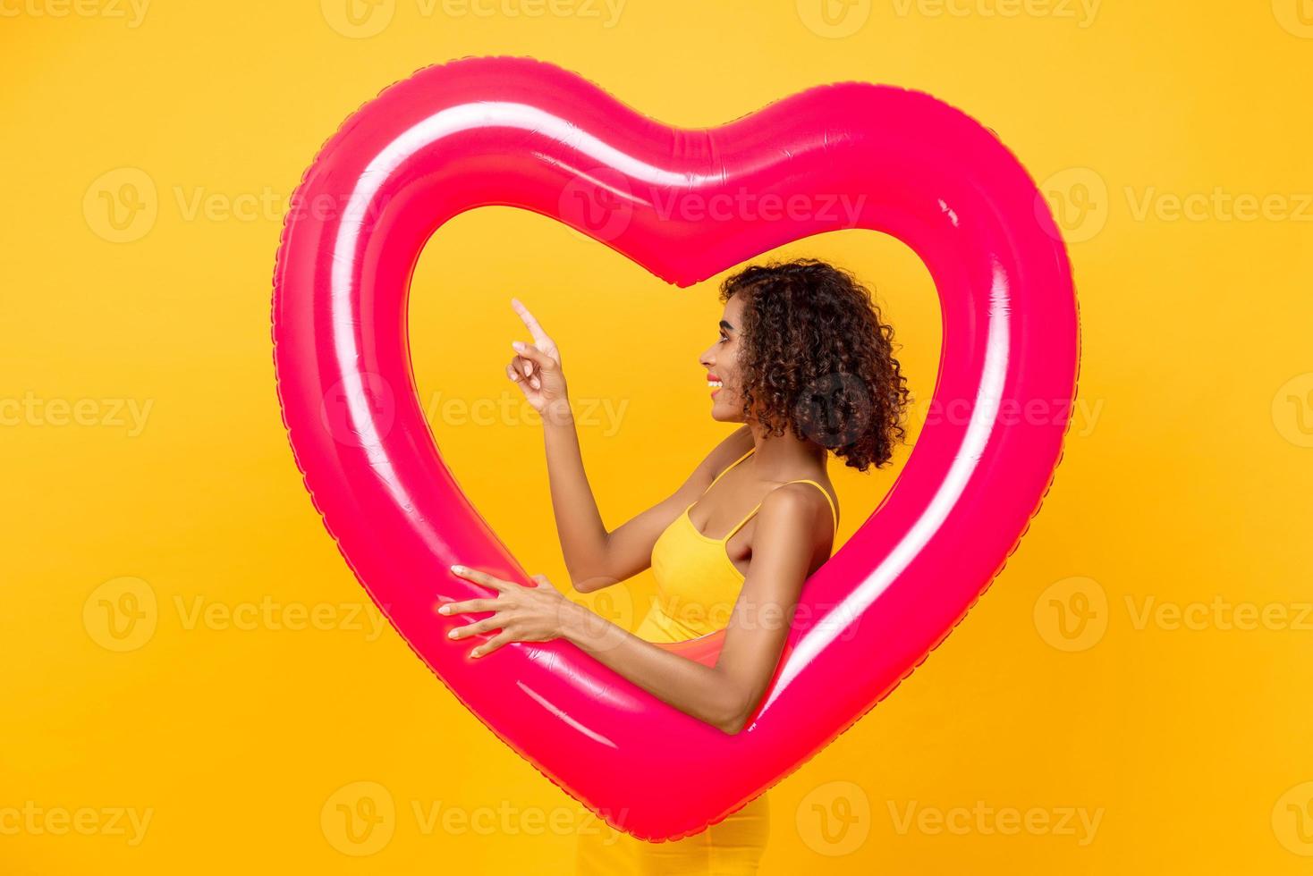 retrato de vista lateral de uma mulher afro-americana sorridente apontando para cima segurando o balão de forma de coração rosa isolado em fundo amarelo foto