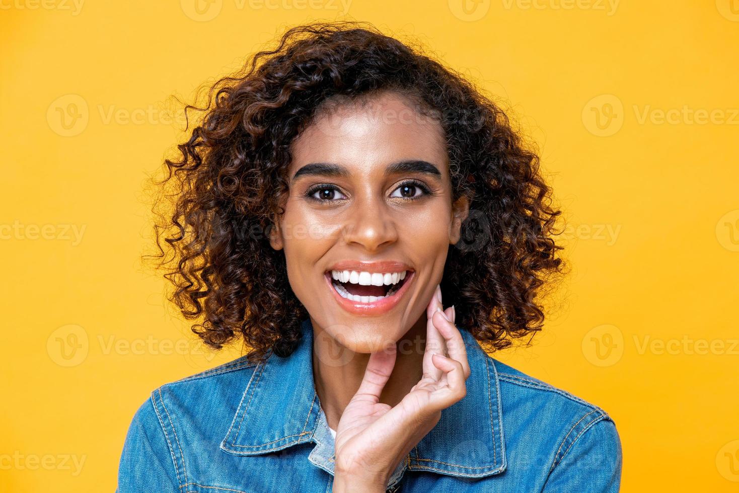 feche o retrato de uma linda e sorridente mulher afro-americana confiante com uma mão tocando seu rosto em fundo de estúdio amarelo isolado foto