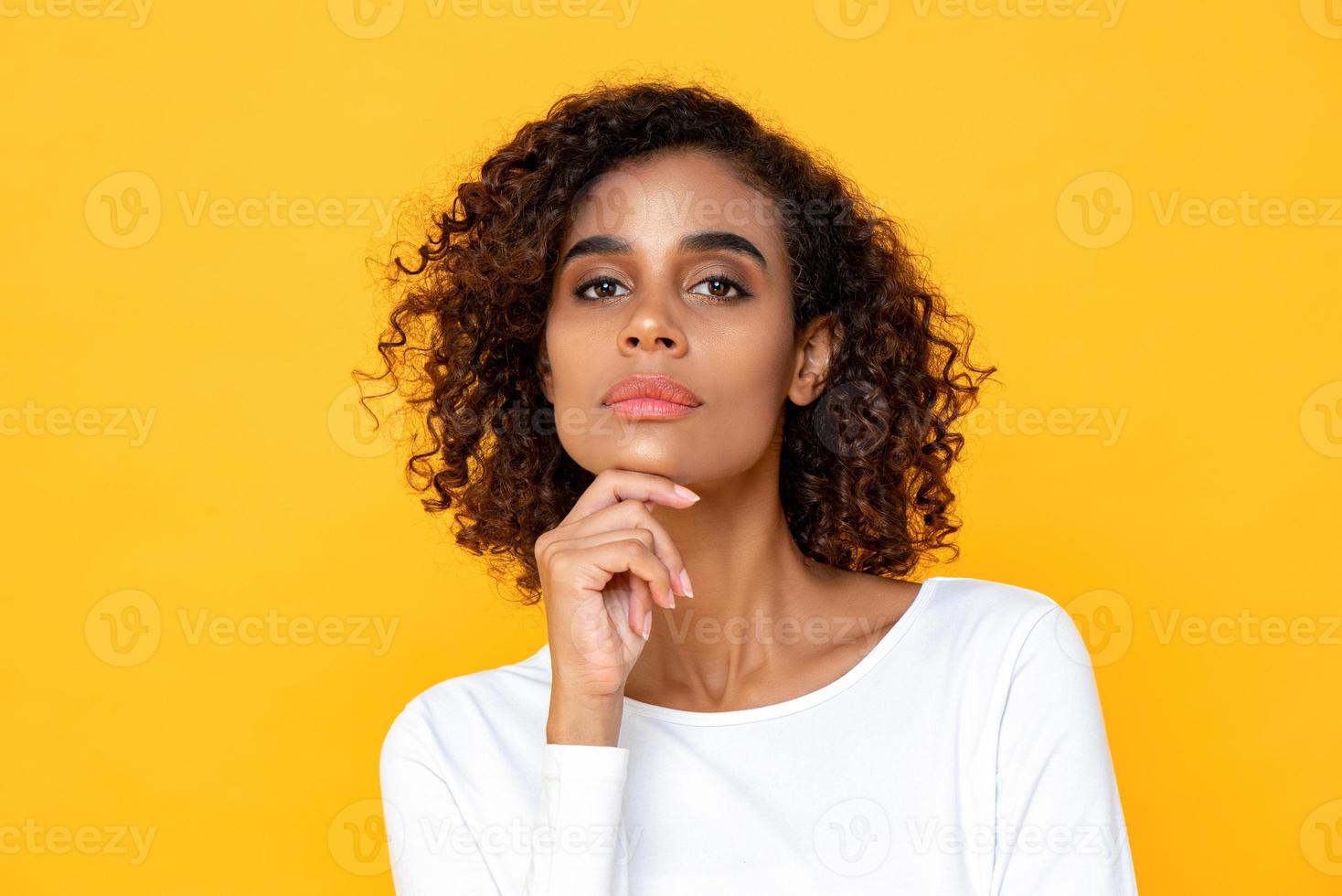 Feche o retrato de pensar jovem afro-americana olhando para a câmera com uma mão tocando o queixo no fundo do estúdio amarelo foto