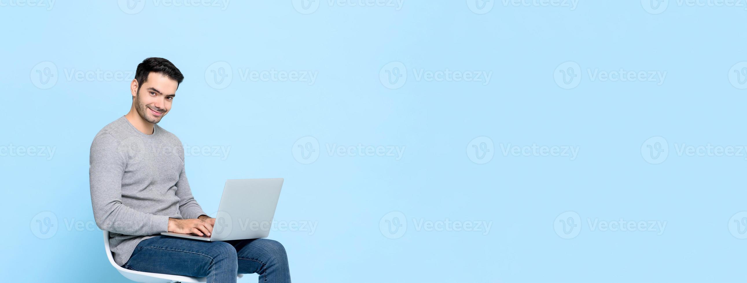 homem bonito sorridente sentado em uma cadeira trabalhando no computador portátil isolado no fundo do banner azul claro com espaço de cópia foto