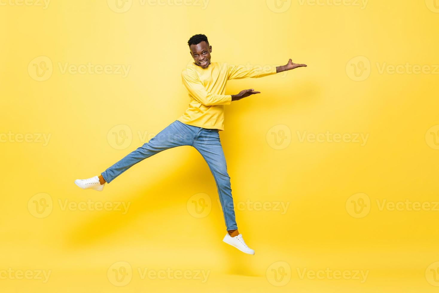 pulando o retrato de um jovem africano enérgico feliz abrindo as mãos para esvaziar o espaço de lado no fundo amarelo isolado do estúdio foto