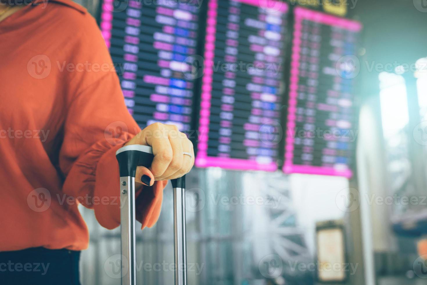 jovem mulher asiática em pé no aeroporto segurando a mala verificando a partida com o horário do voo em segundo plano. foto