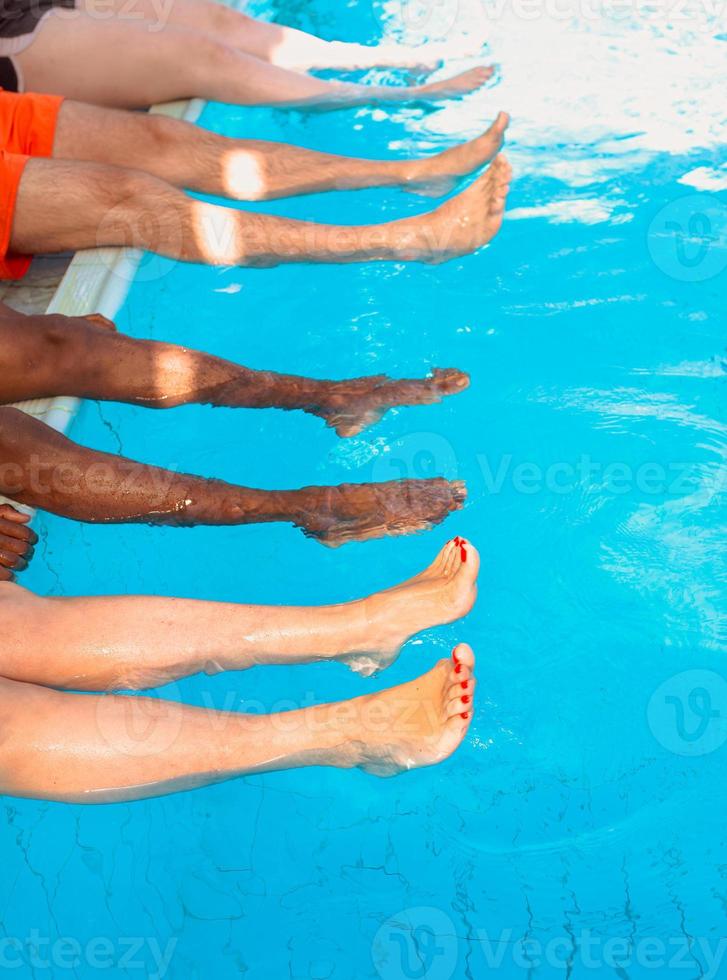 pernas de quatro amigos de raça diferente, sentado à beira da piscina em dia ensolarado. Festa. verão. férias, amizade internacional e conceito de esporte. foto