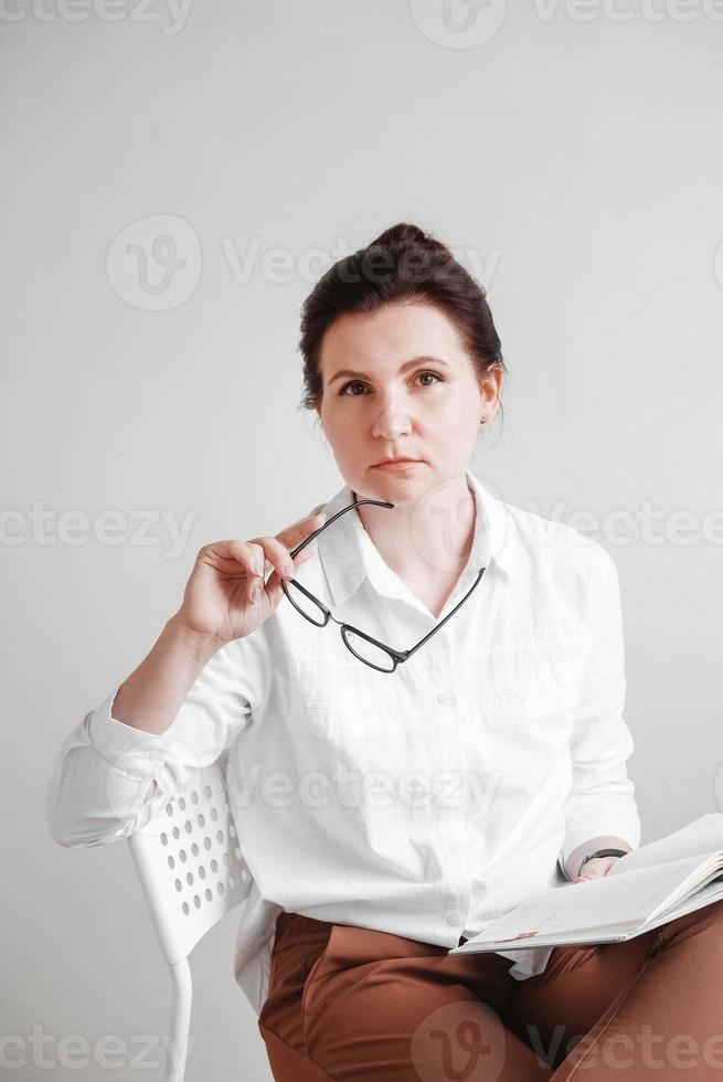 mulher de óculos e vestida com uma camisa branca está sentada em uma cadeira com um livro nas mãos sobre um fundo branco. copiar, espaço vazio foto