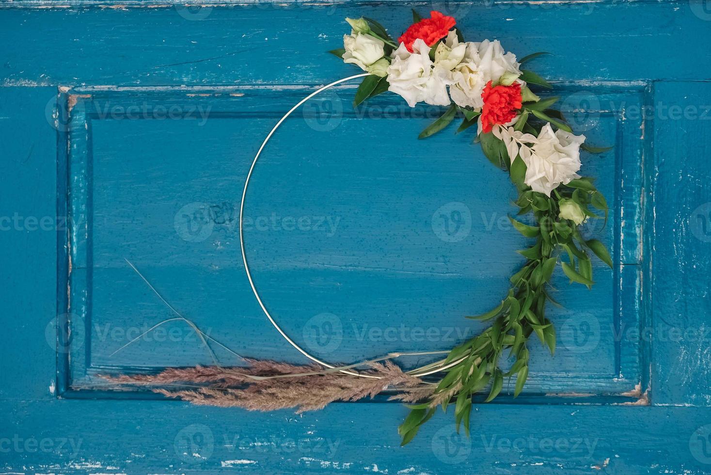 coroa decorada de flores, folhas e grama seca em um fundo de porta azul de madeira. copiar, espaço vazio para texto foto